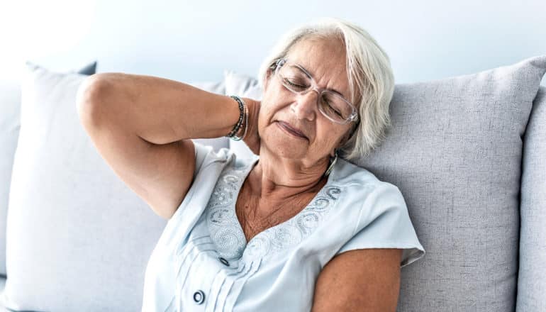 A woman holds the back of her neck while sitting on a couch