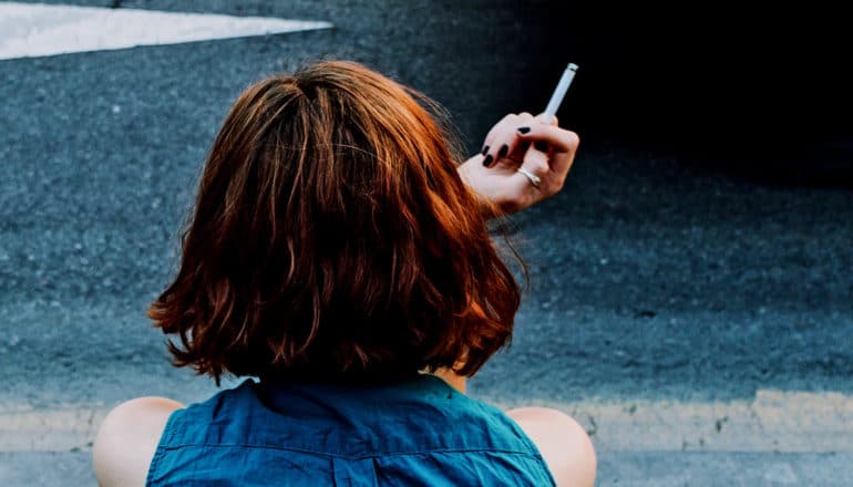 A young woman sits on the sidewalk holding a cigarette