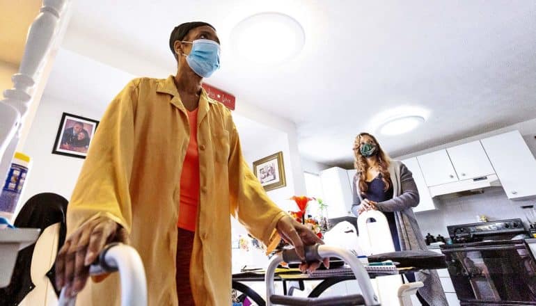 A woman in a yellow house coat stands using a walker while her nurse stands in the background