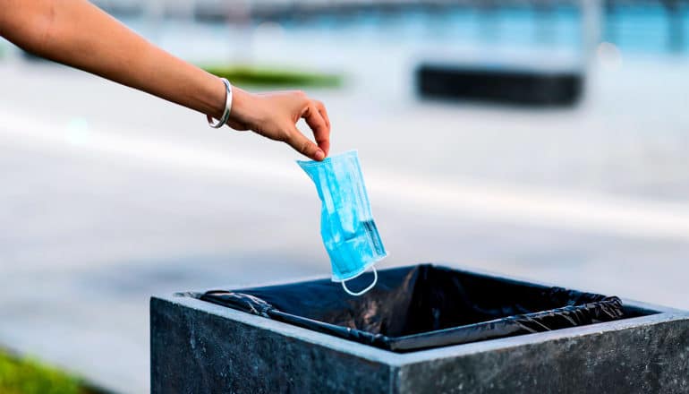 A person drops a blue face mask into a garbage bin