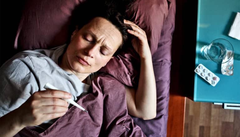 A woman checks her temperature with a thermometer as she lays in bed