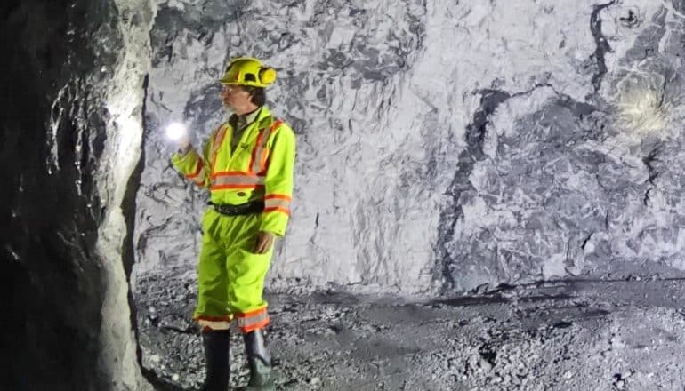 A man in yellow safety gear and a hard hat shines a flashlight at a mine wall