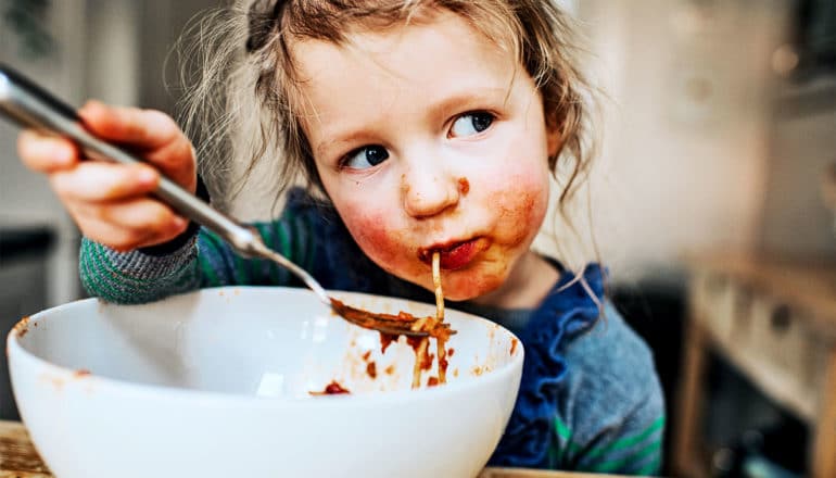 A young girl eats spaghetti from a large bowl, getting sauce all over her face