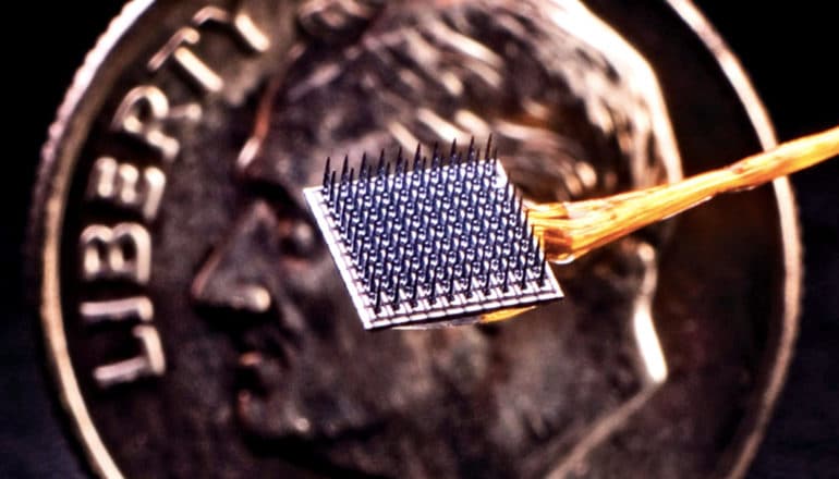 The implant is a small square of metal with spikes, held in front of a penny for scale