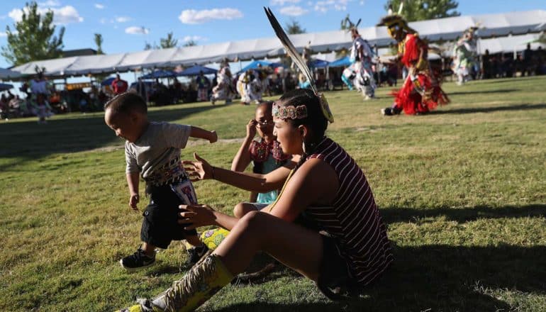 two adults, one in traditional Native American headband with feather and beaded boots, sit outdoors with toddler in front of dancers