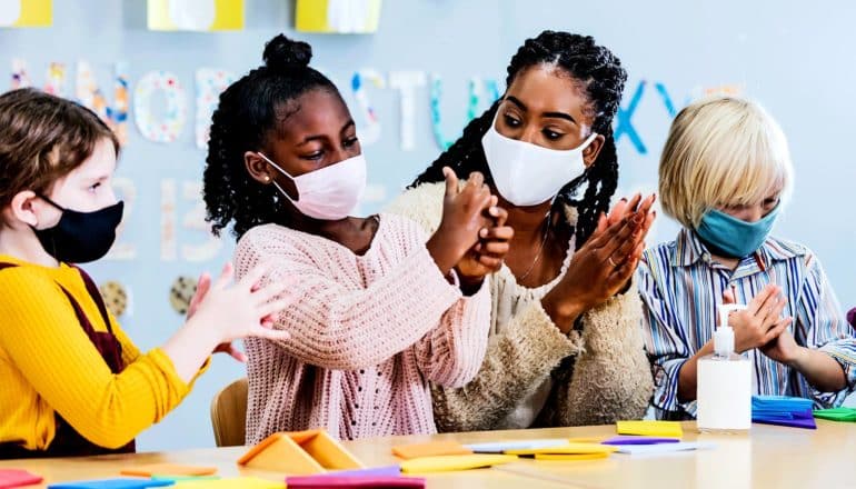 Kids in a classroom use hand sanitizer along with their teacher