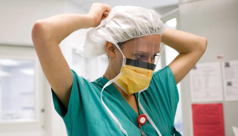 person in scrubs and face shield ties on mask