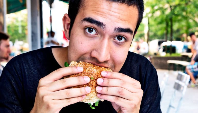 A man eats a burger at a table outdoors while looking up at the camera