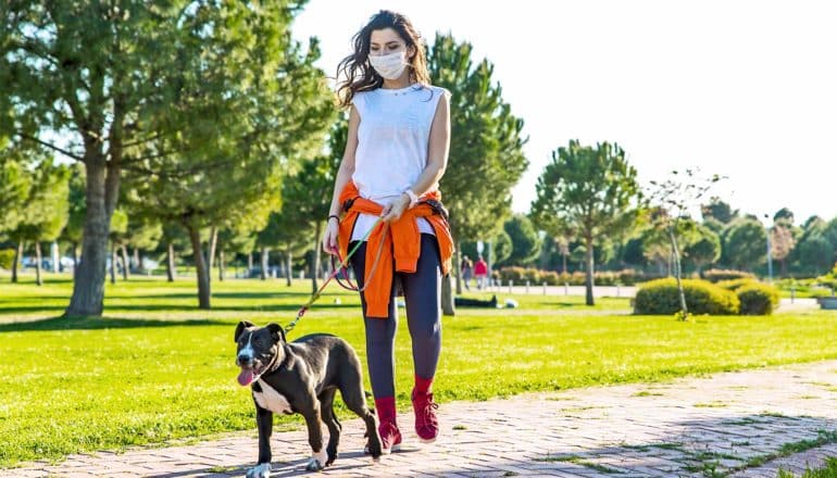 A woman walks her dog through a park while wearing a face mask