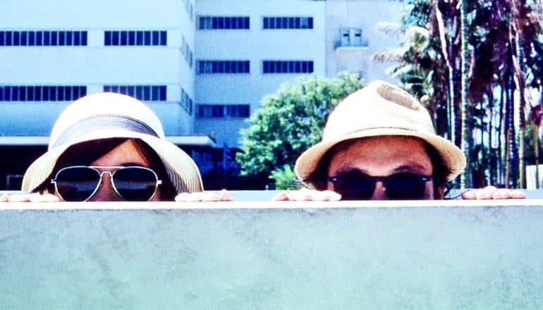 A man and woman in sunglasses and hats peer over a wall