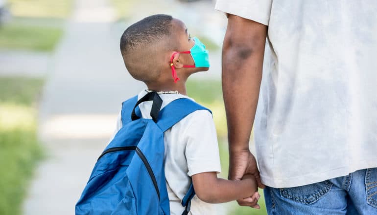 child in mask and backpack looks up at adult and holds hand