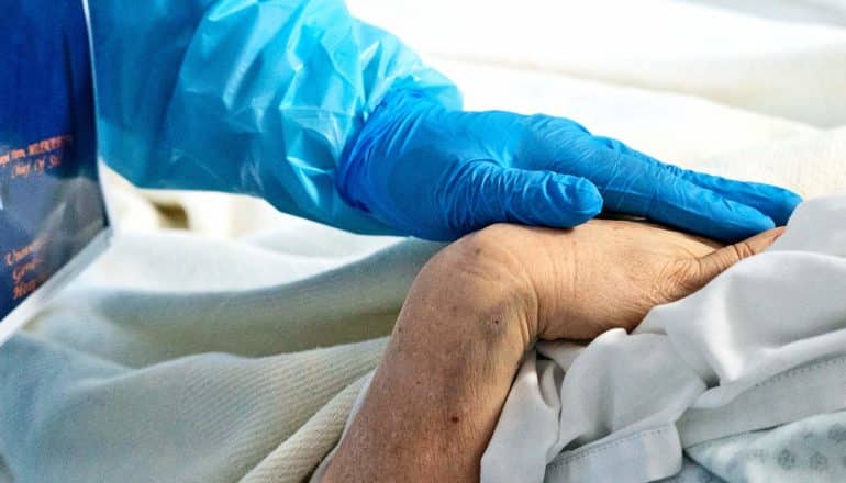 A doctor wearing blue gloves and blue PPE touches a patient's hand