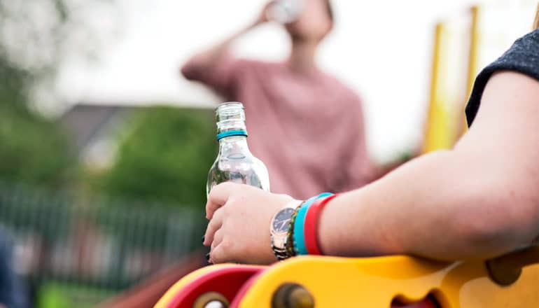 A teenager holds a bottle of alcohol while another sips from a bottle in the background