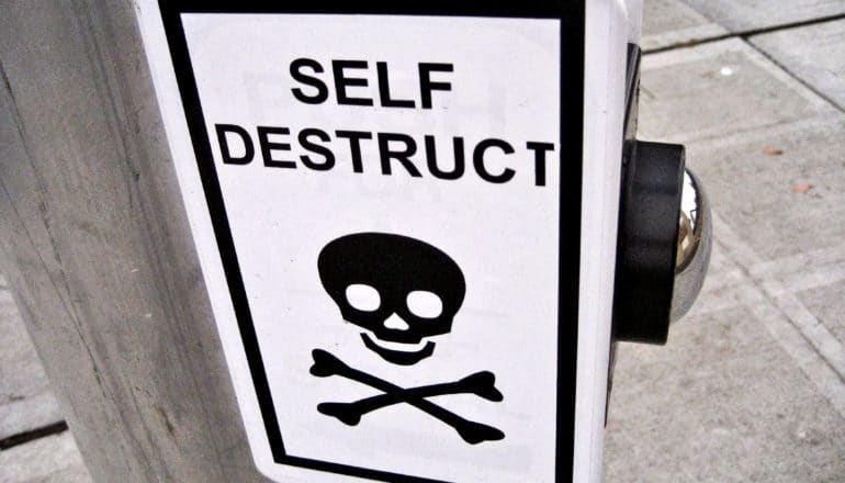 A sign over a button at a crosswalk has a skull and cross bones on it and reads "Self Destruct"