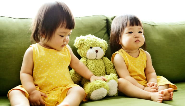 Two twin girls in yellow dresses sit on a couch on either side of a green teddy bear