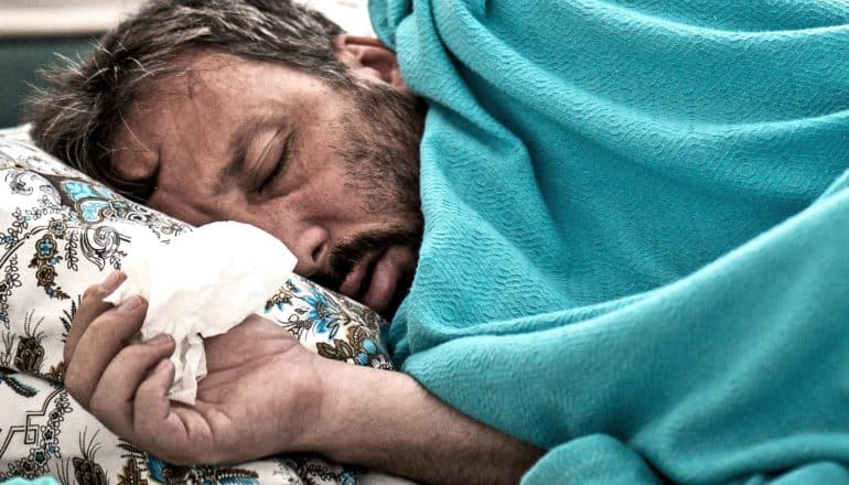 A man holds a tissue while laying under a blue blanket