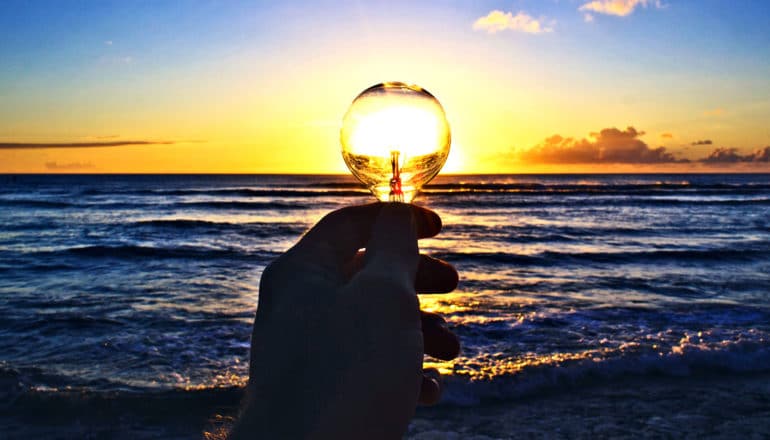 A person holds a lightbulb in front of a sunset over sea water