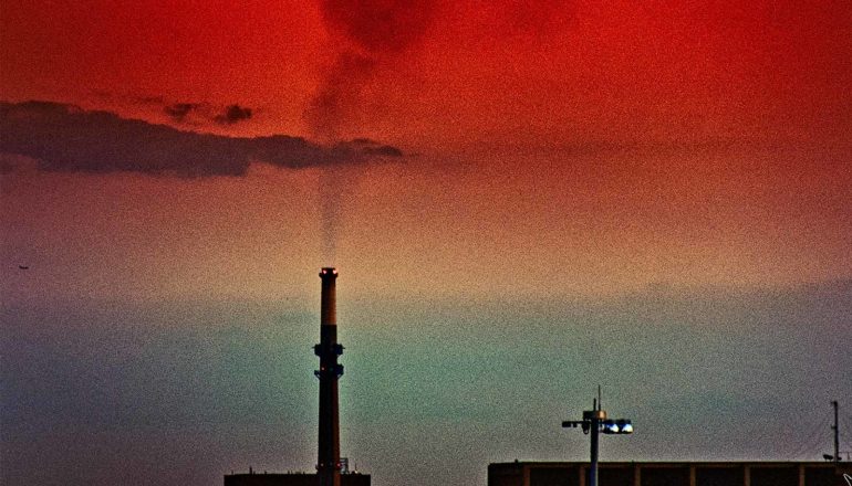 Black smoke rises from a smokestack into a blue, orange, and red sky