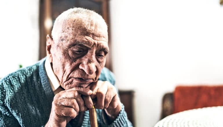 An older man rests his chin on his cane while sitting down at a table