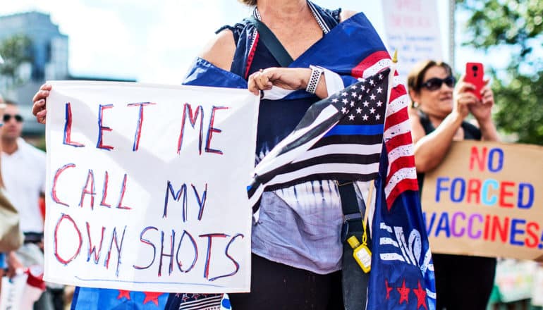 A person wearing several American flags holds a sign that reads "Let Me Call My Own Shots"