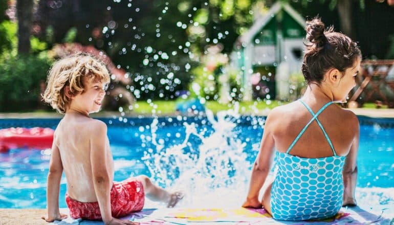 Two kids sit on the side of a pool splashing each other