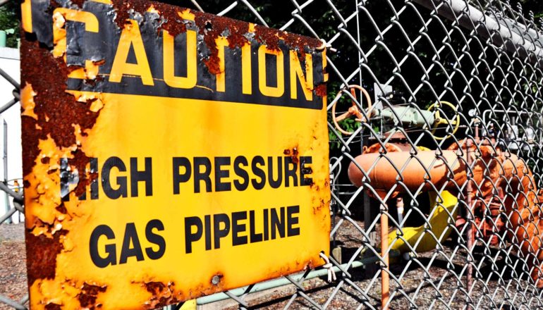 A yellow and black sign on a fence reads "Caution: High Pressure Gas Pipeline"