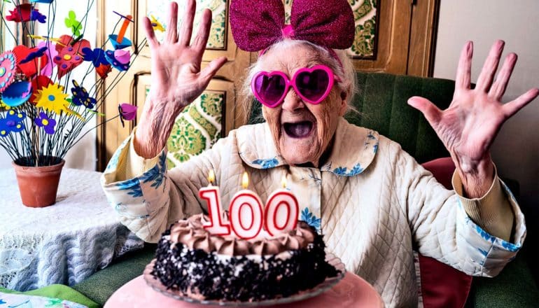 A woman looks excited with pink glasses and a bow on, looking at a cake with "100" in candles