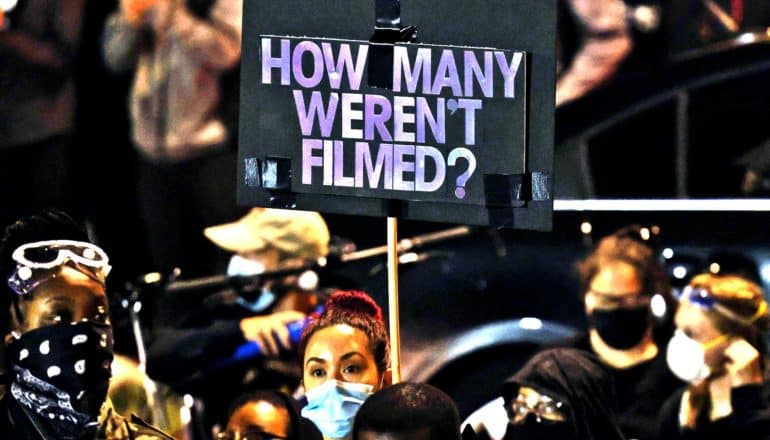 A protester holds a sign that reads "How Many Weren't Filmed?"