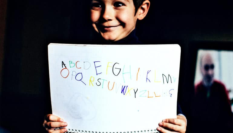 A young boy holds up a sheet of paper with letters written in different colors while smiling