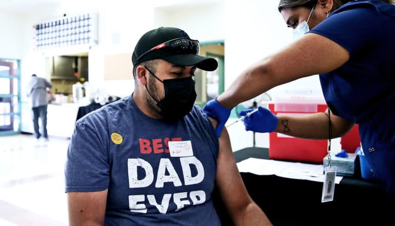 A man wearing a t-shirt that says "Best Dad Ever" gets a COVID-19 vaccine from a health worker