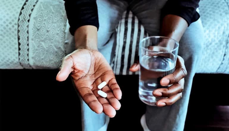 A man sits on the edge of a bed with glass of water and two pills