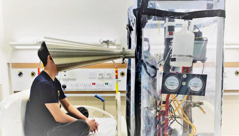 person sits on bed with face in metal cone that leads into plastic-sheathed box of gadgetry