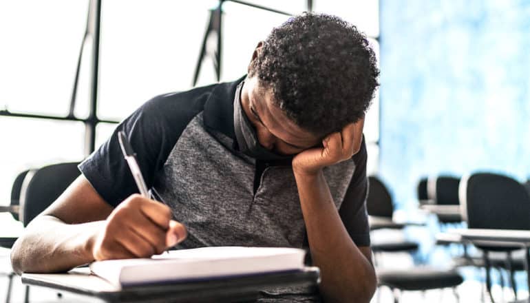 A teen boy sits in class with a mask on and his head resting on his hand as he writes in a notebook