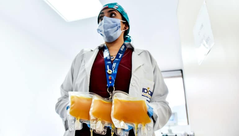 A nurse with a face mask, red scrubs, and a white coat holds three bags of yellow plasma