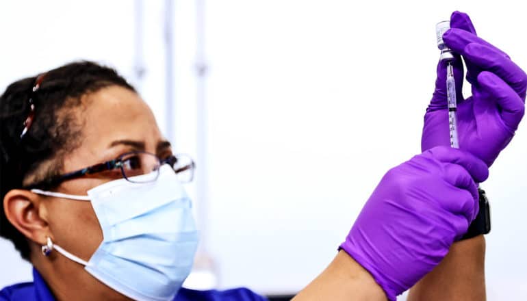 A pharmacist wearing a mask and purple gloves fills a syringe with the COVID-19 vaccine