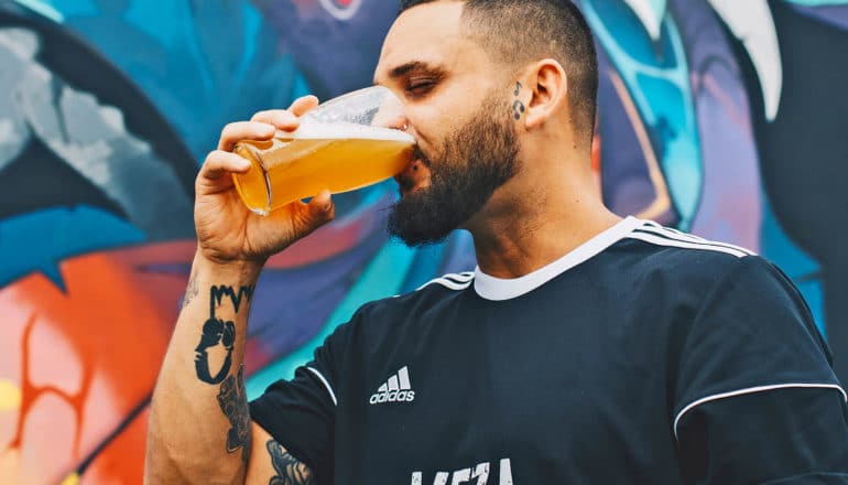 bearded, tattooed person closes eyes while drinking from pint of beer