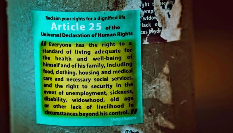 neon sticker on pole says "Reclaim your rights for a dignified life. Article 25 of the Universal Declaration of Human Rights Everyone has the right to a standard of living adequate for the health and well-being of himself and of his family, including food, clothing, housing and medical care and necessary social services, and the right to security in the event of unemployment, sickness, disability, widowhood, old age or other lack of livelihood in circumstances beyond his control. United Nations"