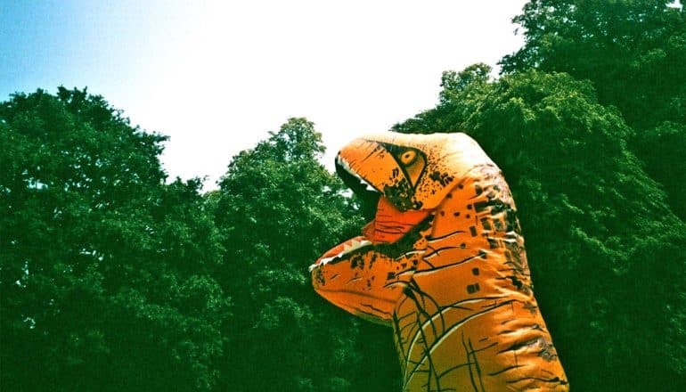 A person in a tyrannosaurus rex costume looks up at some green trees