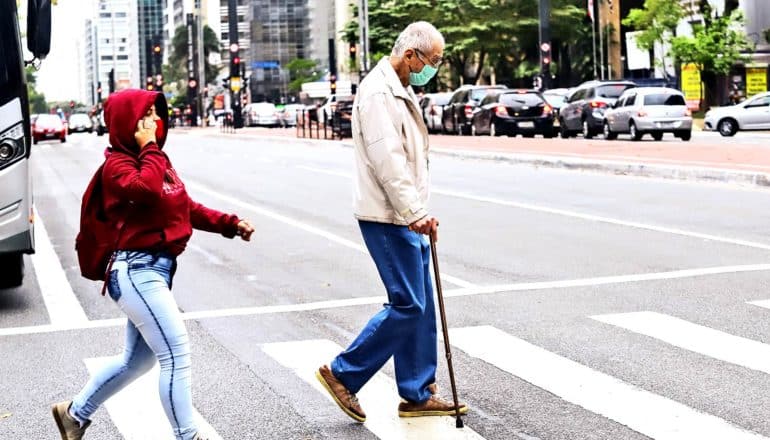 An older man in a face mask walks across a crosswalk with a cane while looking down at the road