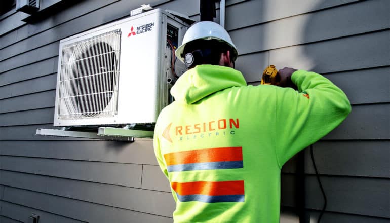 A worker in a bright green hoodie and white hard hat installs a home heat pump outside a house