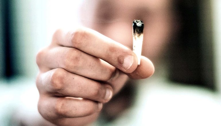 A man holds up a small marijuana joint between his thumb and forefinger