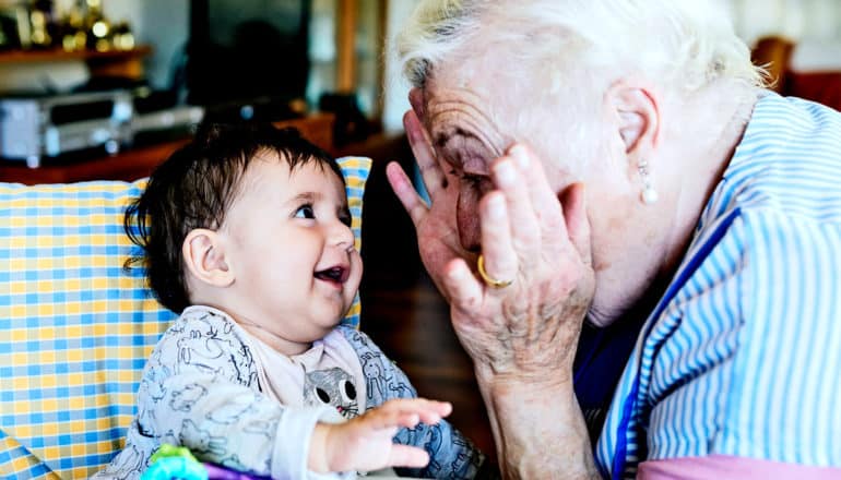 A grandmother plays peek-a-boo with her grandchild, an infant in a high chair