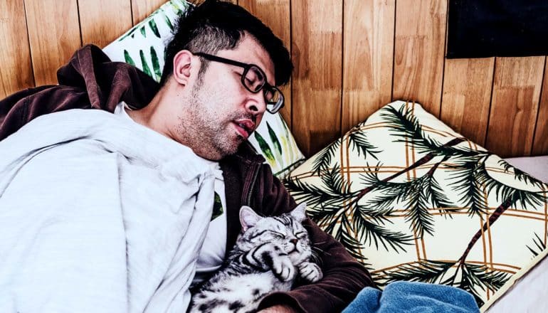 A man naps under a blanket on some pillows against a wood-paneled wall while holding a small cat