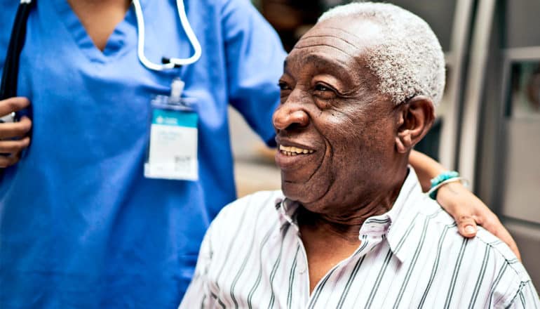 An older man sits while a nurse behind him touches his shoulder