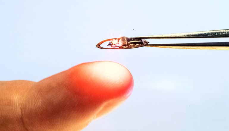 The tiny device is held in tweezers near a person's finger to show it's smaller than a fingertip