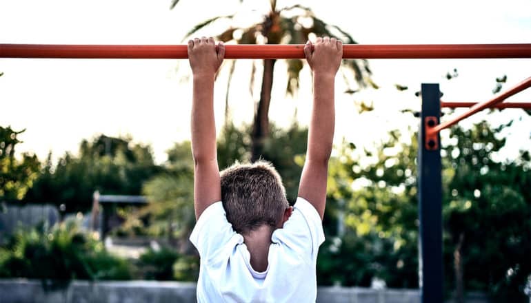 A young boy hangs with both hands from a bar at an outdoor park