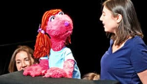 Researchers speak with a pink puppet character with red hair and a blue t-shirt against a black background