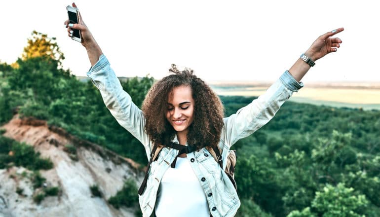 A smiling teen girl in a denim jacket is outside on top of a hill overlooking a forest with her hands in the air