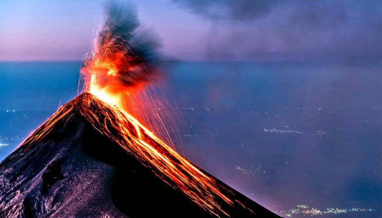 A volcanic eruption at dusk, with red lava flowing down the side of a volcano