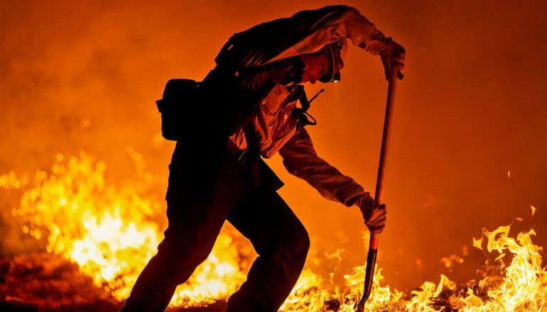 A firefighter uses a shovel to secure a fire break while flames burn behind him and the air is bright orange